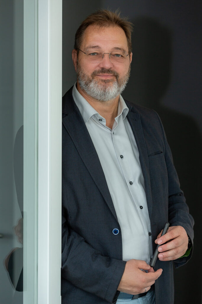 Mittelalterlicher Mann mit grauem Bart und Brille lächelt freundlich in dunklem Anzug